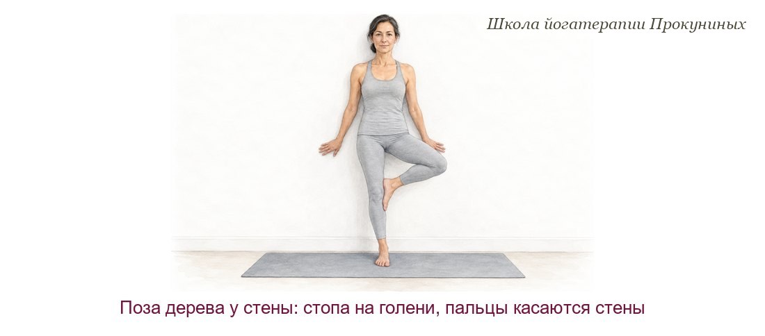 Женщина стоит в позе дерева спиной у стены: стопа на голени опорной ноги, кончики пальцев касаются стены